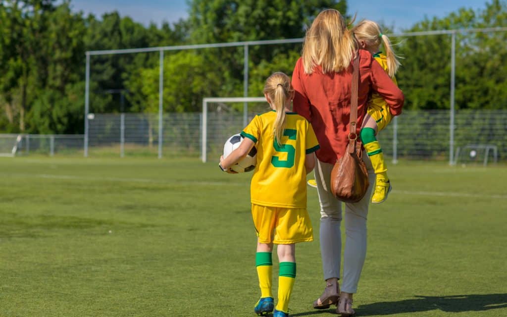 Mutter geht mit zwei Töchtern im Fußballtrikot über den Sportplatz, Symbol dafür, wie Eltern ihre Kinder im Sport begleiten und unterstützen.