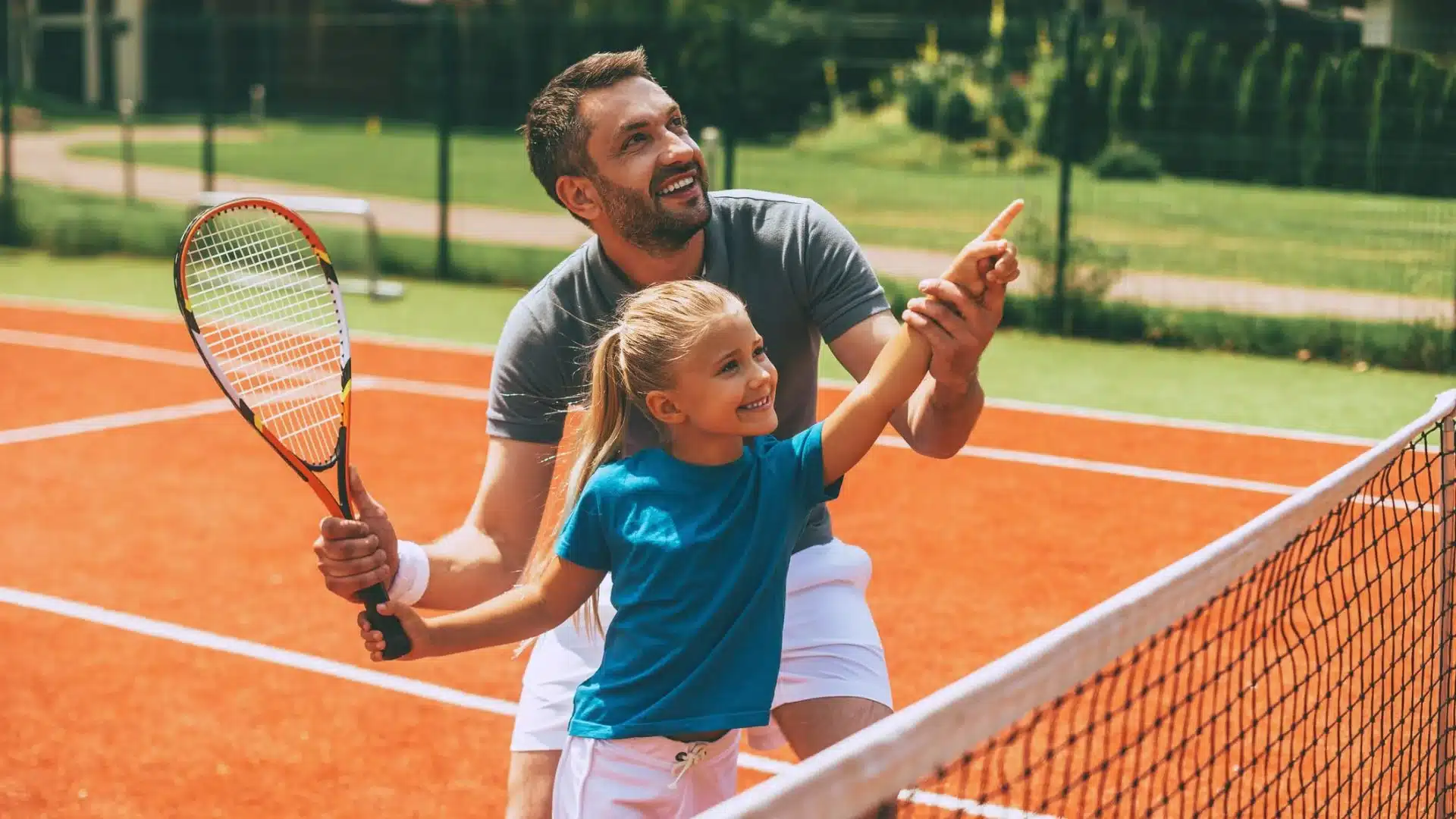 Vater spielt mit seiner Tochter Tennis und zeigt ihr lächelnd die Schlagrichtung – Symbol dafür, wie Eltern ihre Kinder im Sport positiv begleiten und Selbstvertrauen stärken.
