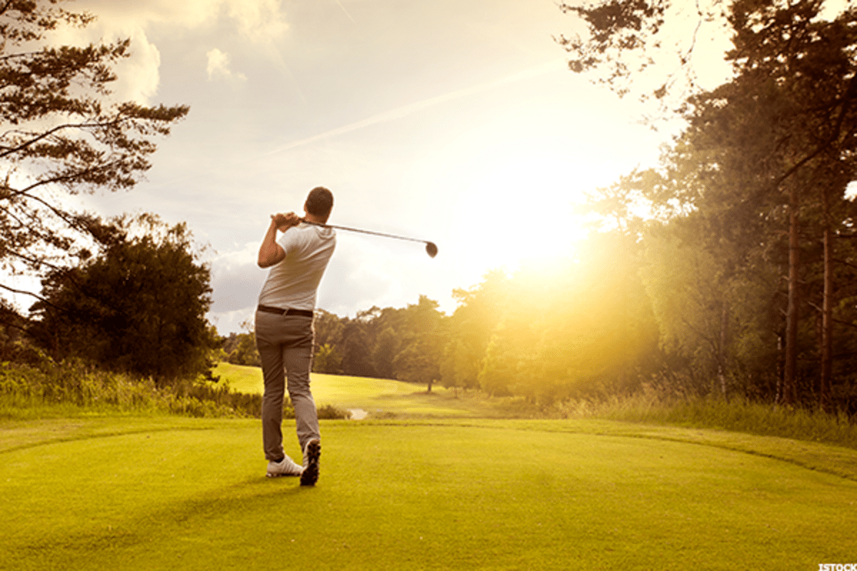 Golfer beim vollen Schwung auf einem hellen, grünen Fairway bei Sonnenuntergang, Fokus auf Dynamik und Weite des Platzes.