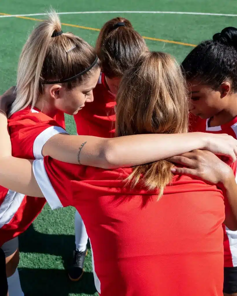 Fußballerinnen im Teamkreis mit verschränkten Armen auf dem Spielfeld – Symbol für Zusammenhalt, Teamgeist und gemeinsame mentale Wettkampfvorbereitung von Teams und Vereinen.