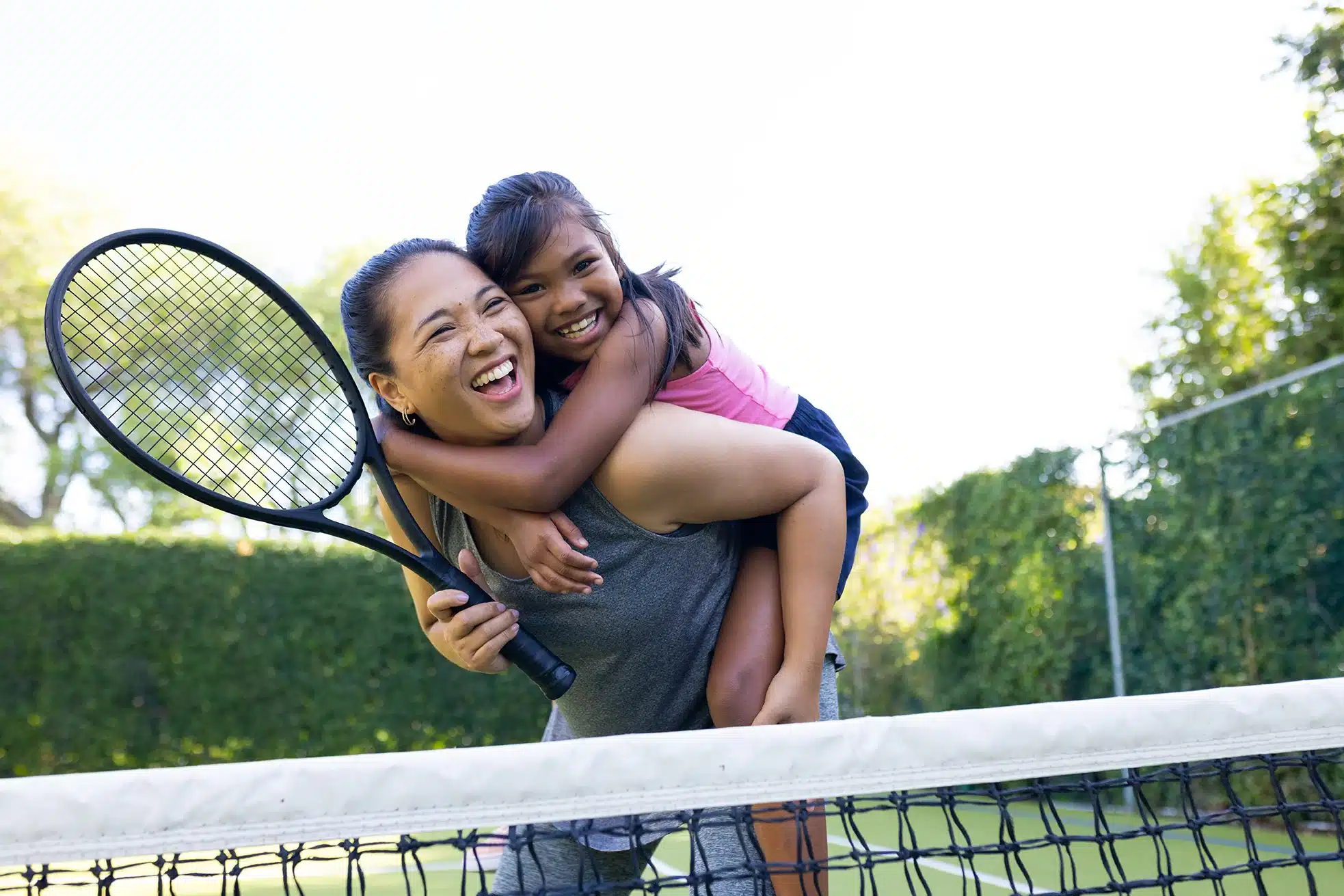 Mutter mit Tennisschläger trägt lachend ihre Tochter auf dem Rücken auf dem Tennisplatz – zeigt, wie Eltern ihre Kinder im Sport liebevoll unterstützen und Freude am Sport fördern.